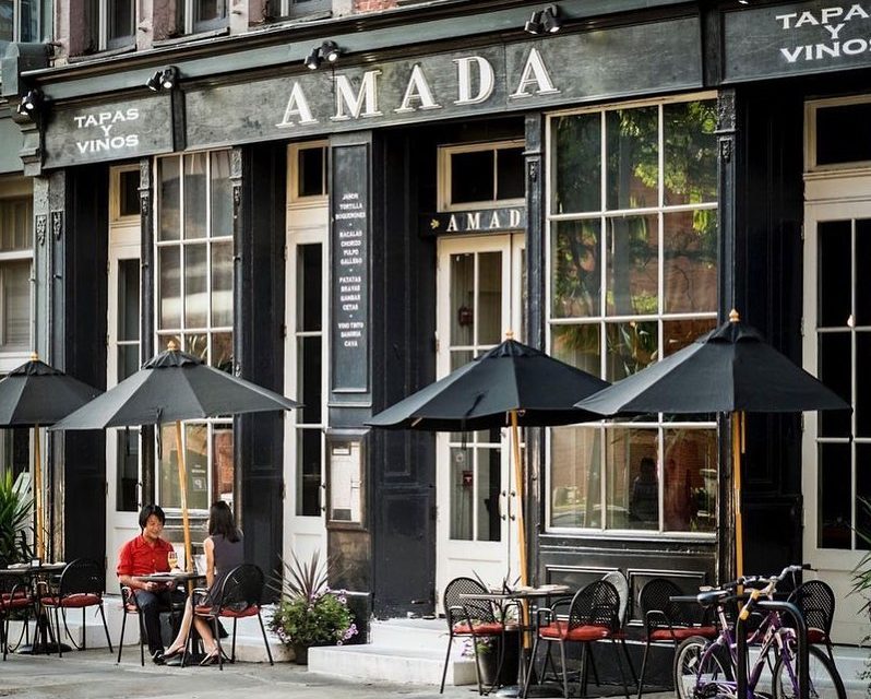 Front of Amada restaurant with tables and umbrellas on the sidewalk and a couple dining.