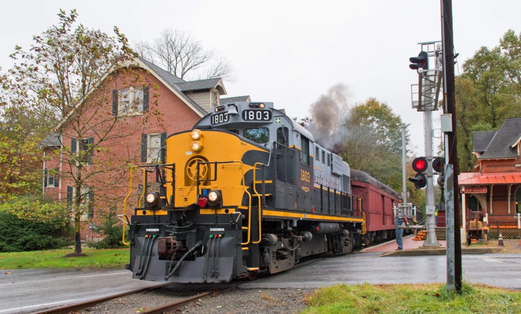 A West Chester Railroad locomotive with a bright yellow front stopped at a brick train station.