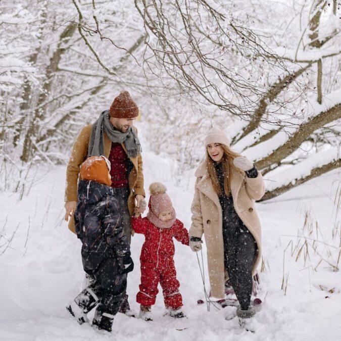 A mother and father and two young children wear winter coats and snow gear while walking in the snowy woods.