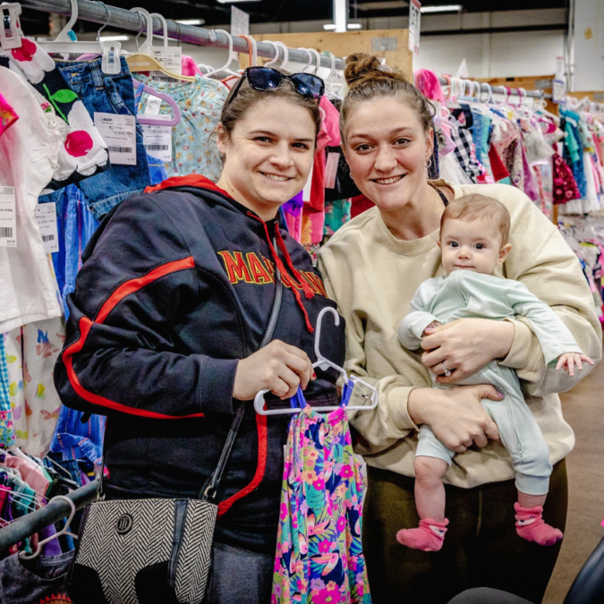 Two women, on holding a baby are shopping for resale baby gear
