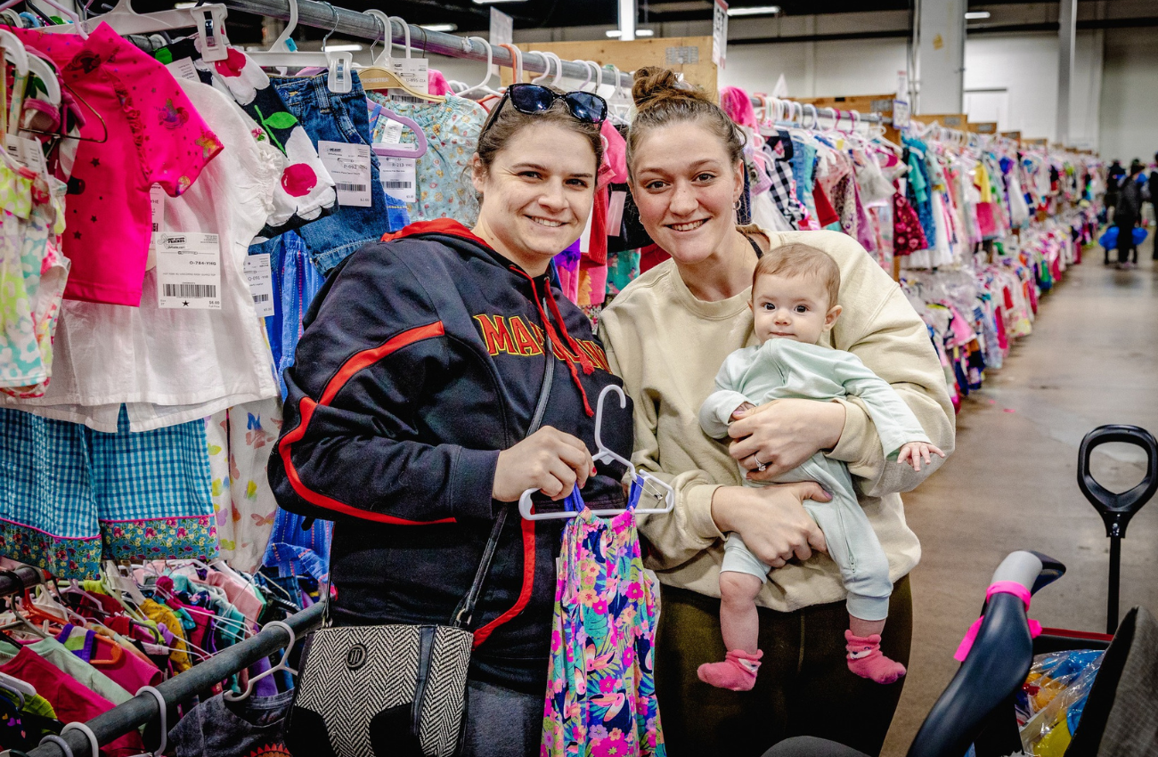 Two women, on holding a baby are shopping for resale baby gear