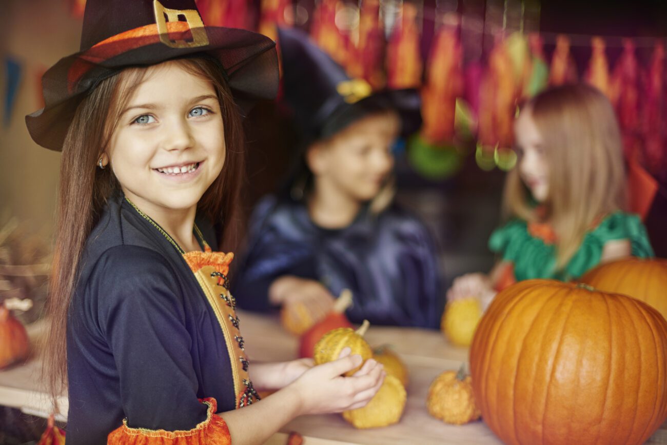 Young girl in a witch costume next to pumpkins with two other little girls in costume in the background.