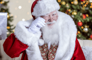 Santa winks his eye in a red suit with a Christmas tree in the background.