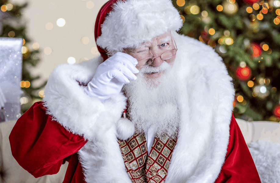 Santa winks his eye in a red suit with a Christmas tree in the background.