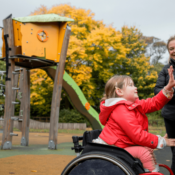 Girl in red jacket in a wheelchair plays on an accessible playground with her mother.