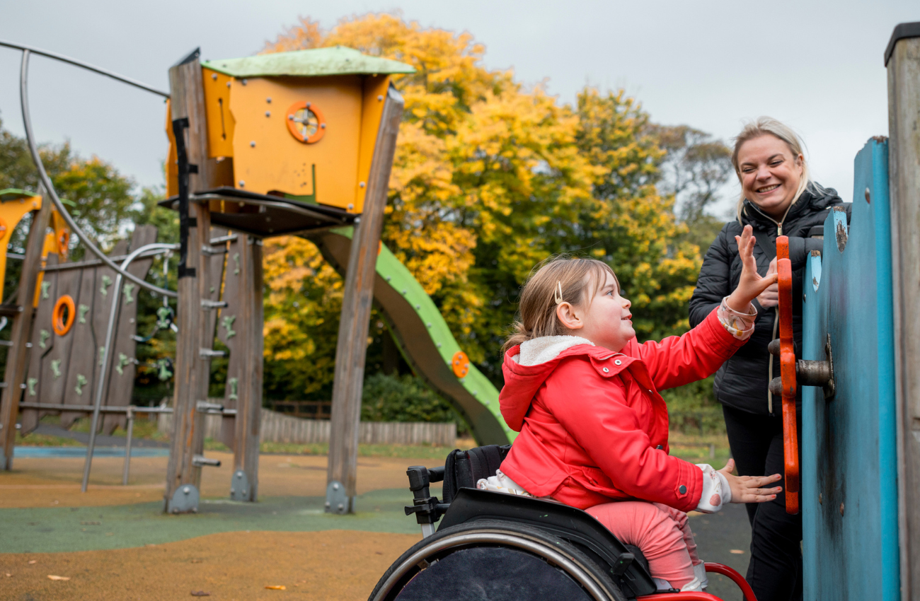 Girl in red jacket in a wheelchair plays on an accessible playground with her mother.
