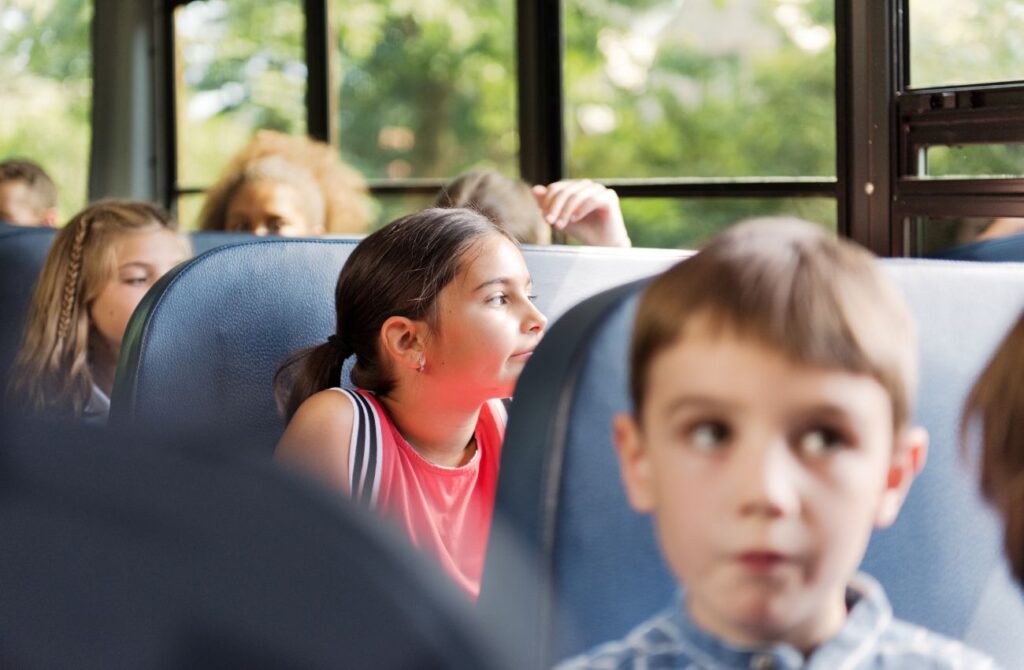 A young school girl looks out the window of a full school bus with an ambivalent expression.  