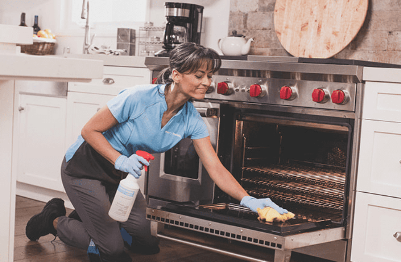 A woman in a blue shirt performs house cleaning services and wipes the inside of an oven.