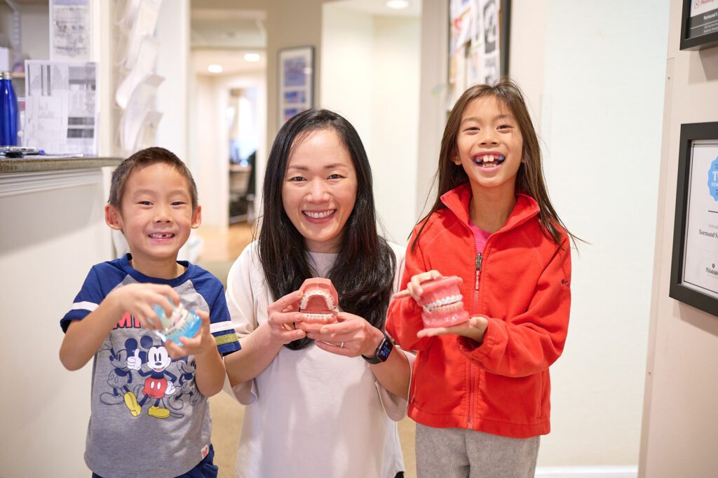 Orthodontist Dr Yang posing with two children each holding a fake set of teeth and smiling