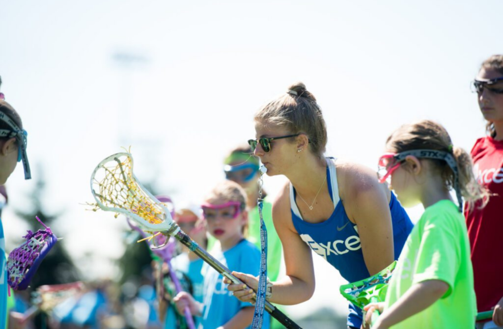 A teen girl displays leadership skills, one of many summer camp benefits, while demonstrating lacrosse skills to younger girls.