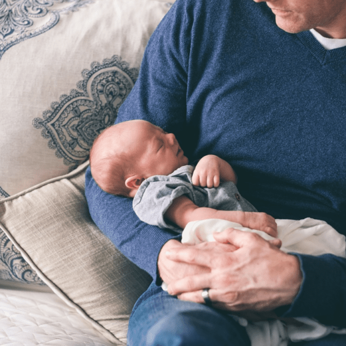 A new dad on paternity leave holds his baby with his wife looking over his shoulder.