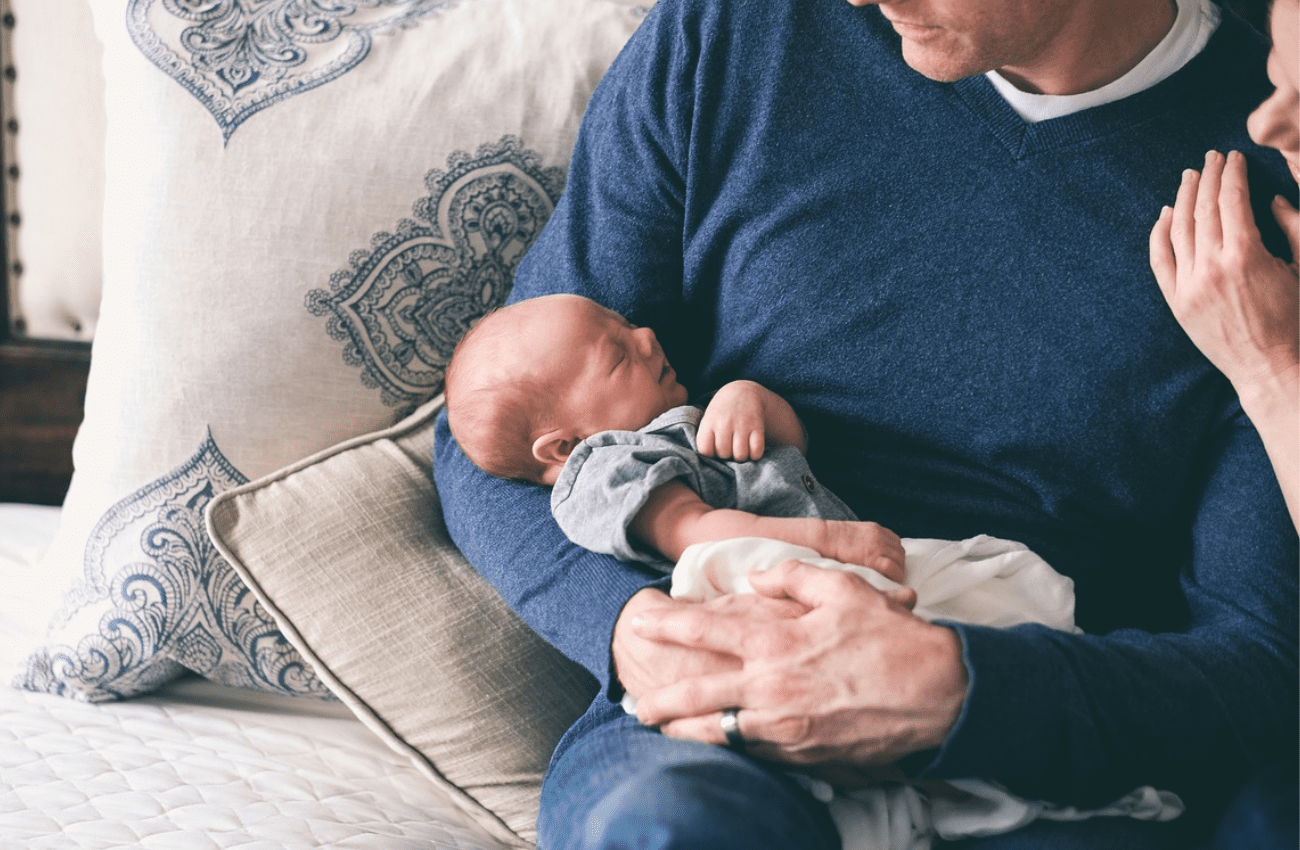 A new dad on paternity leave holds his baby with his wife looking over his shoulder.