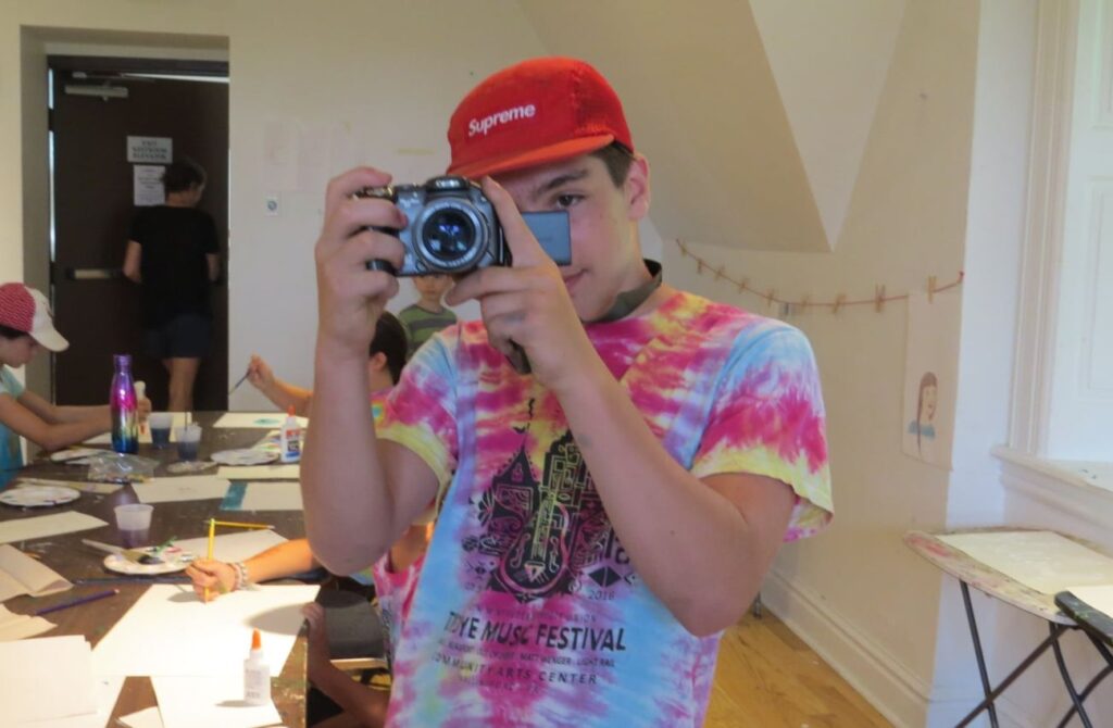 A teen boy points a digital camera at the viewer in a youth arts class at Community Arts Center.
