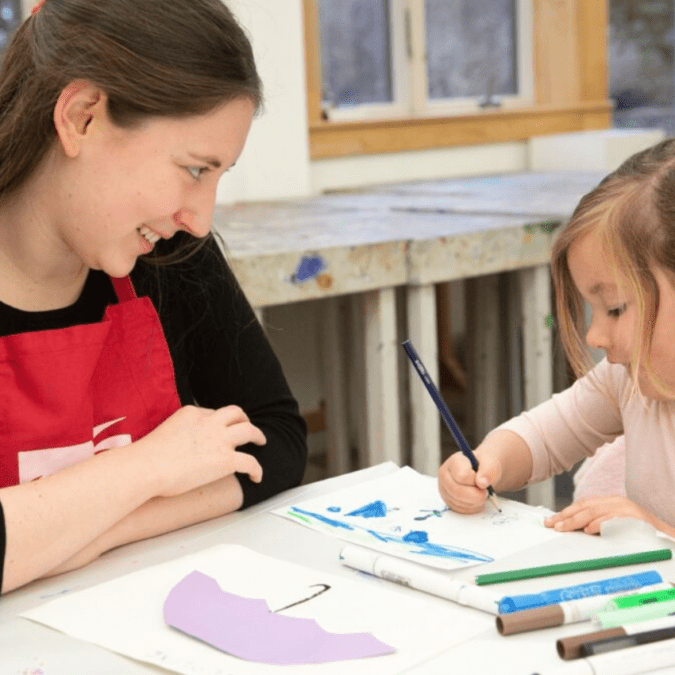 A young woman art teacher smiles at a little girl drawing in one of the art classes for kids at Wayne Art Center.