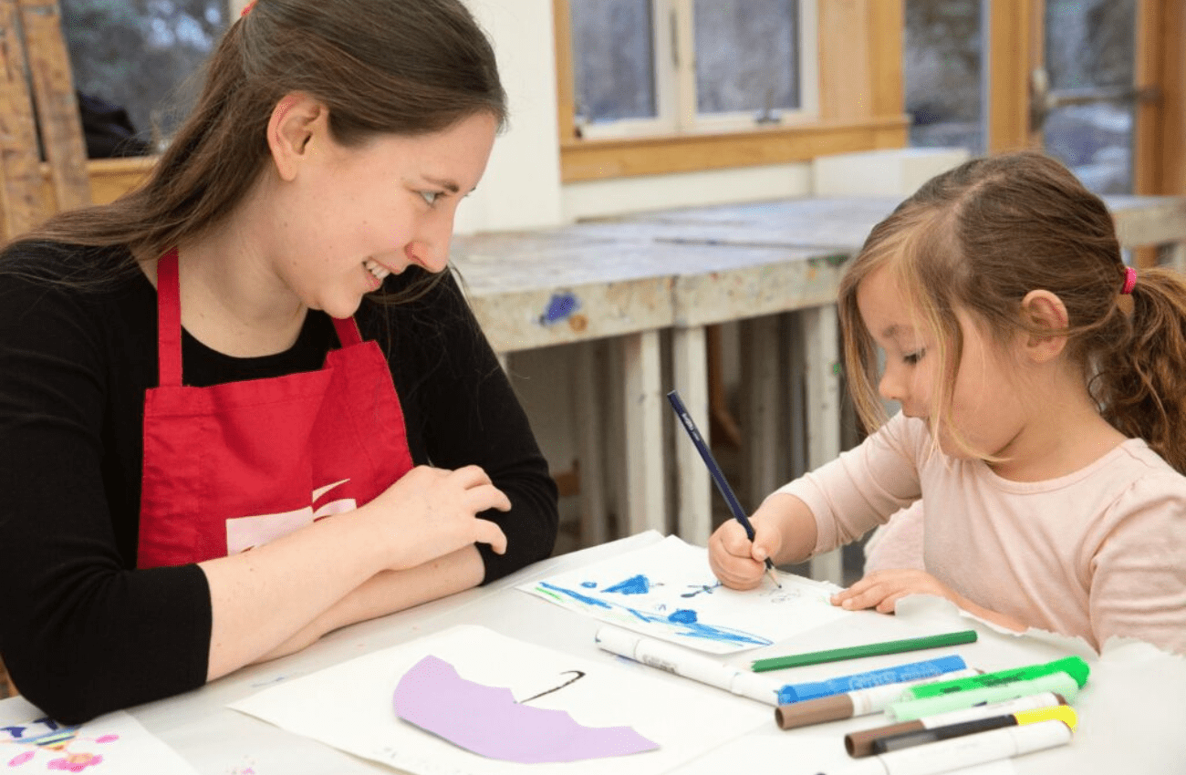 A young woman art teacher smiles at a little girl drawing in one of the art classes for kids at Wayne Art Center.