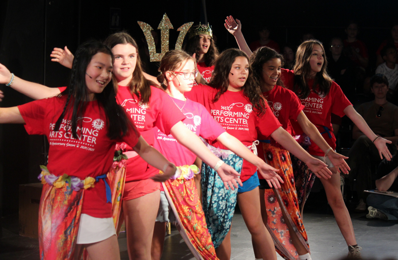 Performers on stage at Wolf Performing Arts Center, children are wearing red shirts with brown vests.