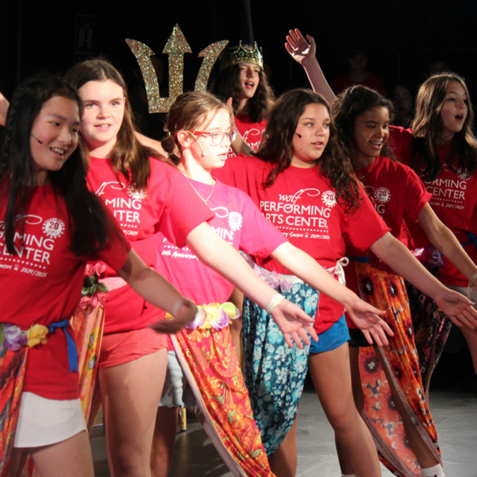 Performers on stage at Wolf Performing Arts Center, children are wearing red shirts with brown vests.