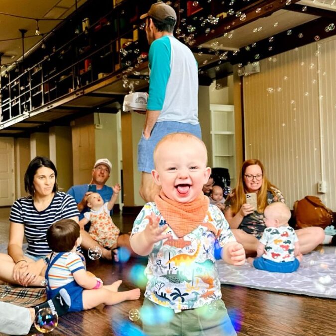 A walking baby laughs for the camera with bubbles and moms and babies in the background during a music class for babies.