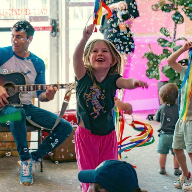 Mister John's Music plays guitar at a kids party with bubles and rainbow pom-poms.