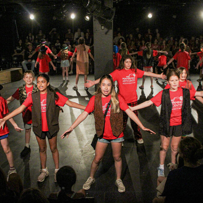 Performers on stage at Wolf Performing Arts Center, children are wearing red shirts with brown vests.