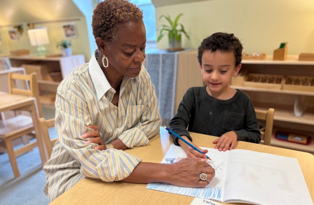 A preschool boy hold a pencil above his drawing while his teacher guides him.