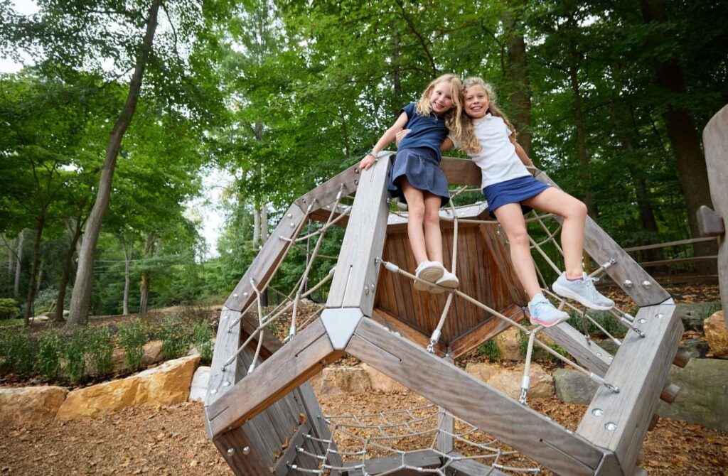 Two little girls smile while perched on a climbing cube in a wooded playground at Benchmark School. 