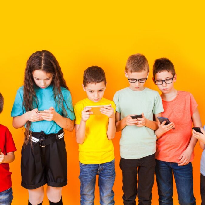 Six elementary school kids look at screens against a yellow wall.