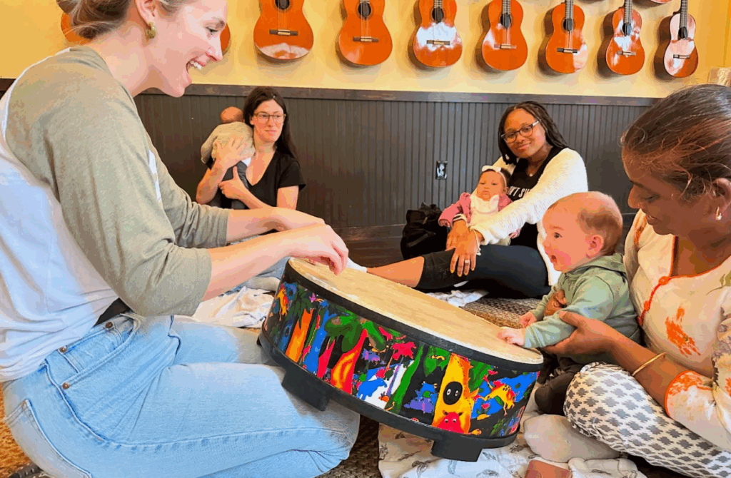 A baby and teacher both touch a coloful drum in a Mister John's Music baby and mom music class. 