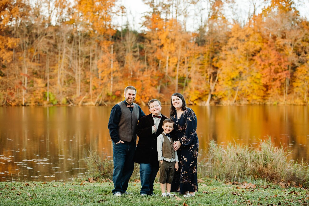 The Thomas family with their two young boys in front of a lake with fall foliage