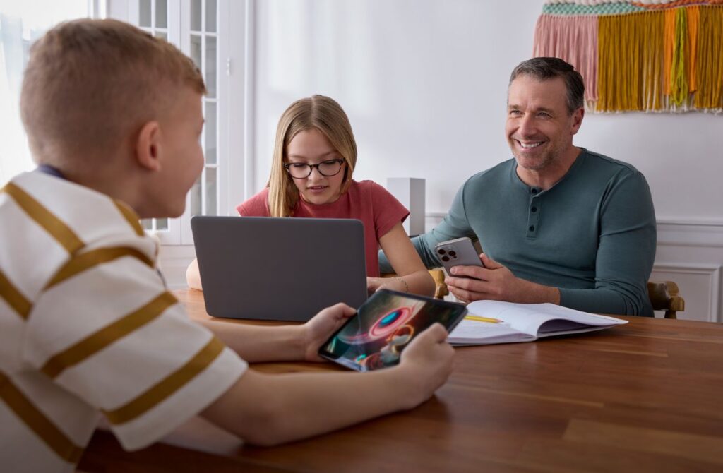 A father, teen daughter, and pre-teen son sit around a table on their devices using Xfinity connectivity. 