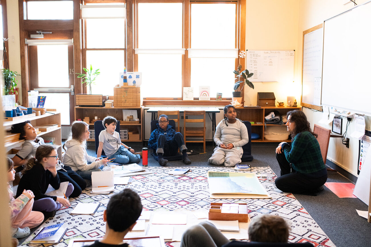 Students at Gladwyne Montessori sitting with their teacher on the carpet while learning a lesson, these are middle school students.