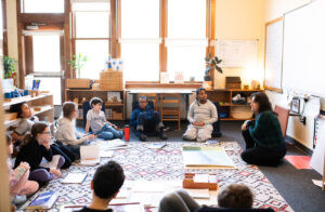 Students at Gladwyne Montessori sitting with their teacher on the carpet while learning a lesson, these are middle school students.