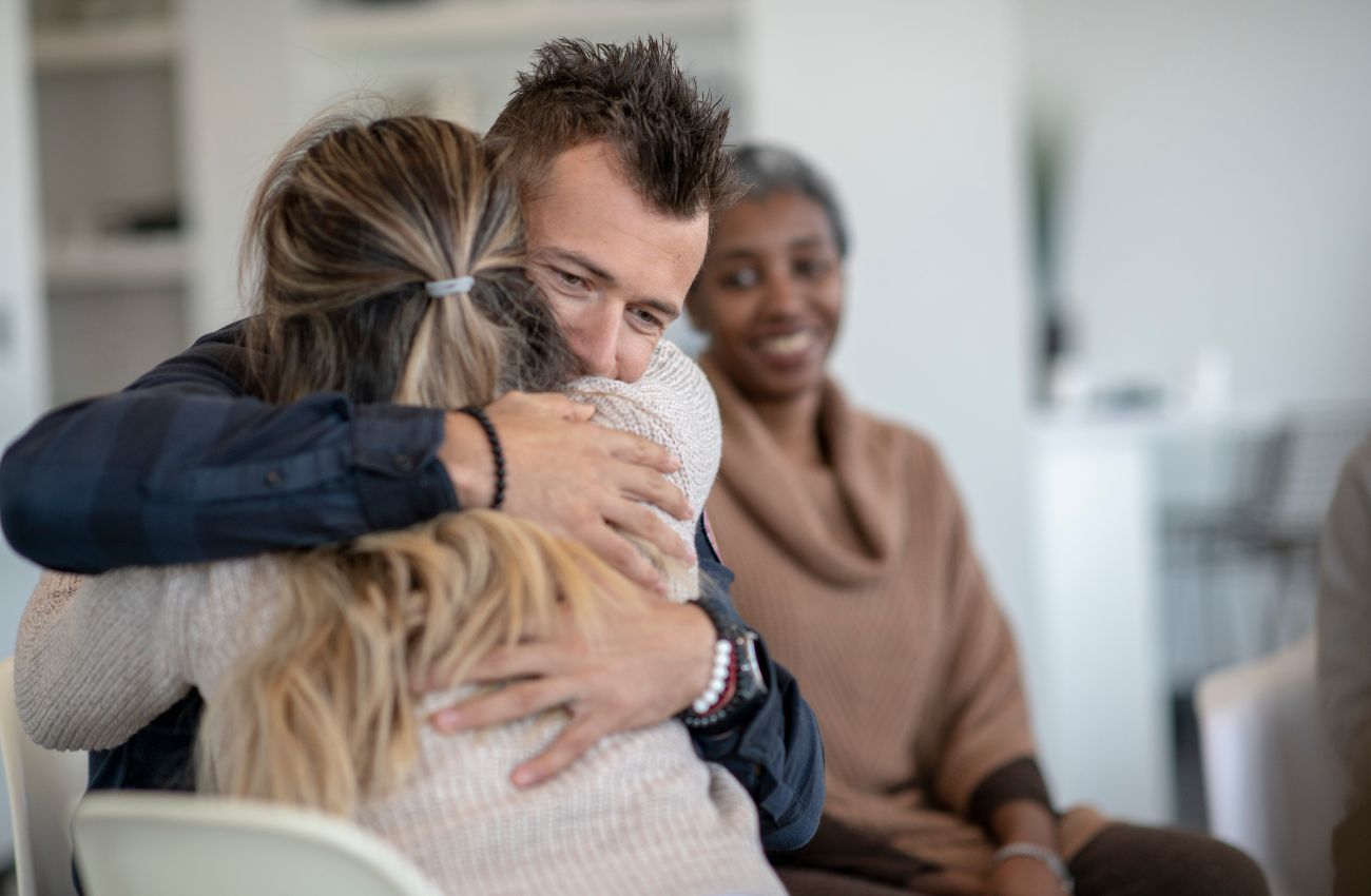 Support from friends, family, and professionals are important elements for recovery from alcohol dependency. A man and woman in recovery hug while another woman looks on.