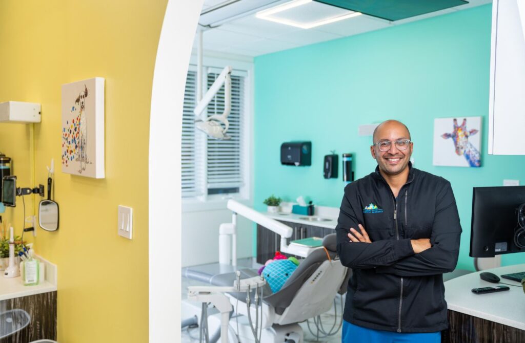 Dr. Anil Shah stands in in front of an aqua blue exam room of his pediatric dentistry office.