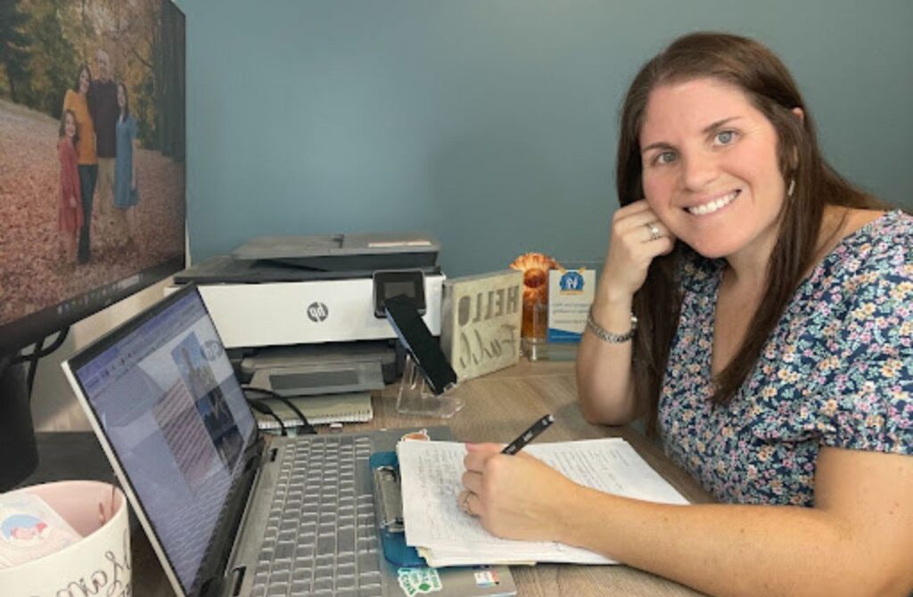 Pediatric sleep trainer, Katie Roeder sits at her desk writing notes with her laptop open.