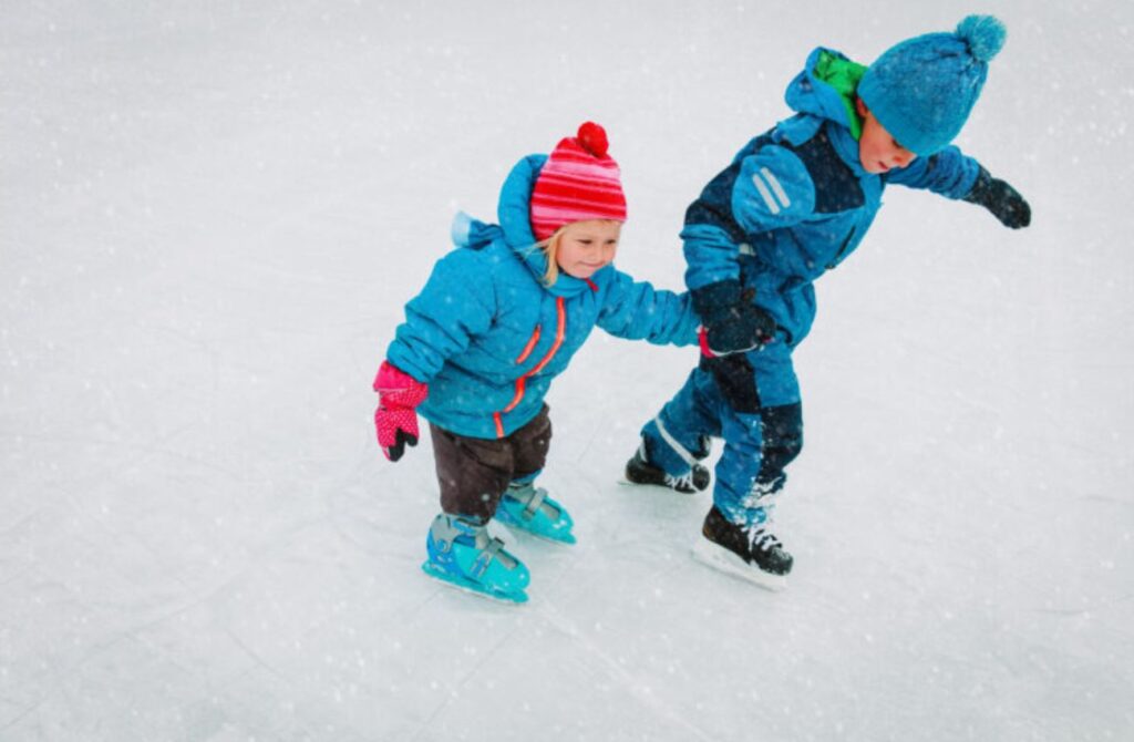 Bundled up for winter, a toddler girl and little boy hold hands while ice skating outdoors. 