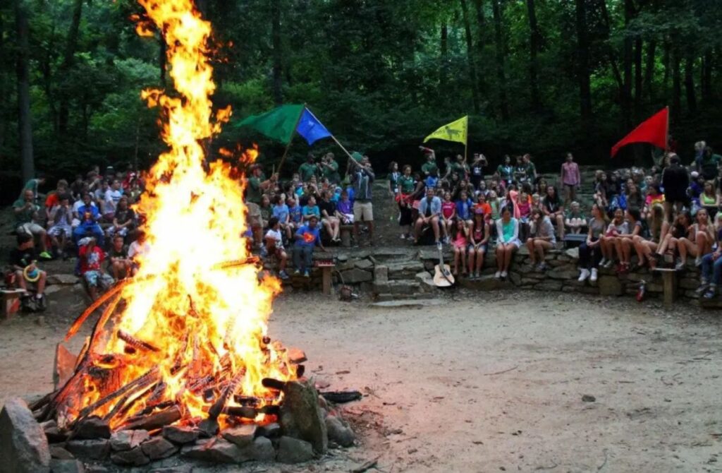 Four colored flags wave as a huge daytime bonfire burns surrounded by large numbers of campers at Camp Conrad Weiser. 