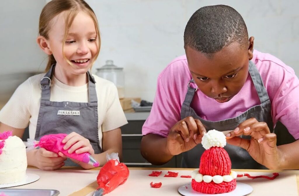 A young boy in a pink tee shirt and apron carefully ices a winter-hat-shaped confection while a young girl in an apron watches at a culinary day off camp.