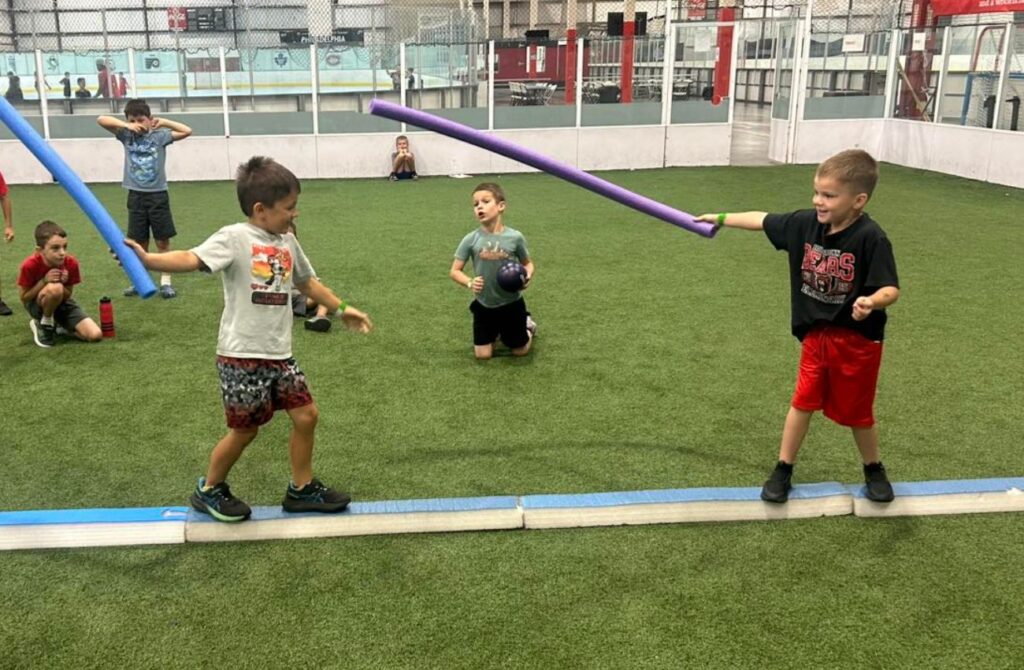 Two boys have a foam tube duel while balancing on a foam beam while other day camp kids watch.