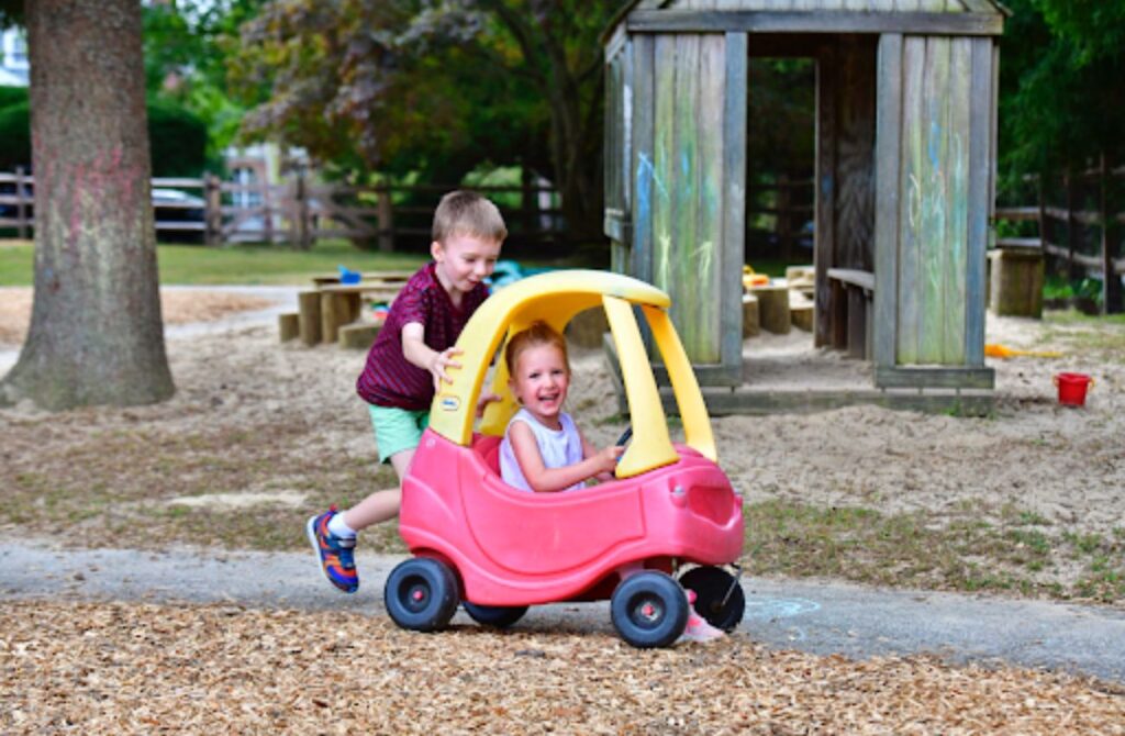 A little boy pushes a little girl in a red and yellow rideable car outdoors at Phebe Anna Thorne School in Bryn Mawr. 
