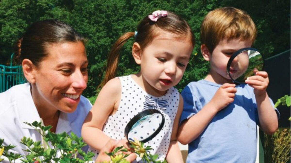 Preschool-aged children and an adult gaze at plants with a magnifying glass.