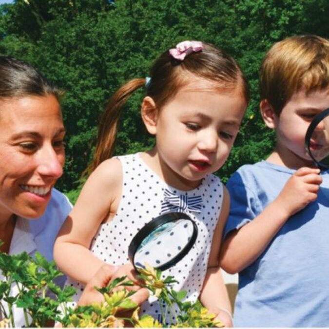 Preschool-aged children and an adult gaze at plants with a magnifying glass.