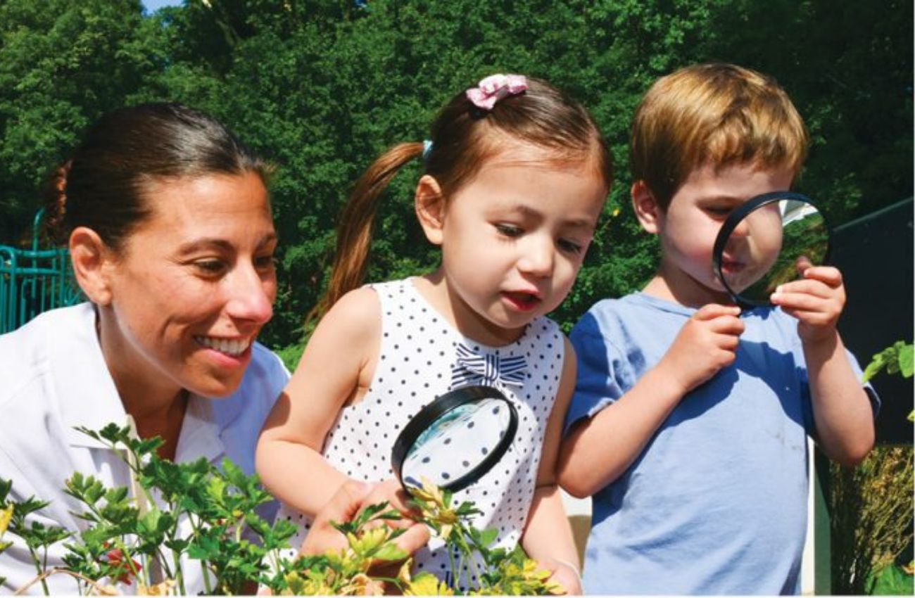 Preschool-aged children and an adult gaze at plants with a magnifying glass.