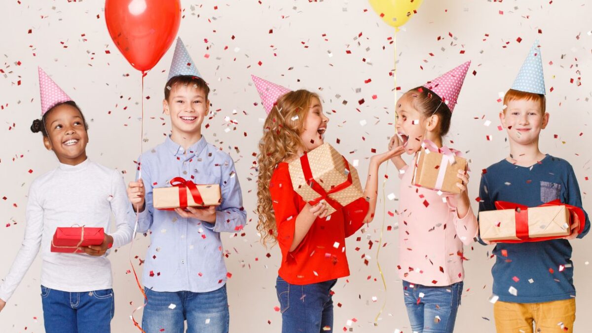 Five kids in party hats hold presents at a birthday party.