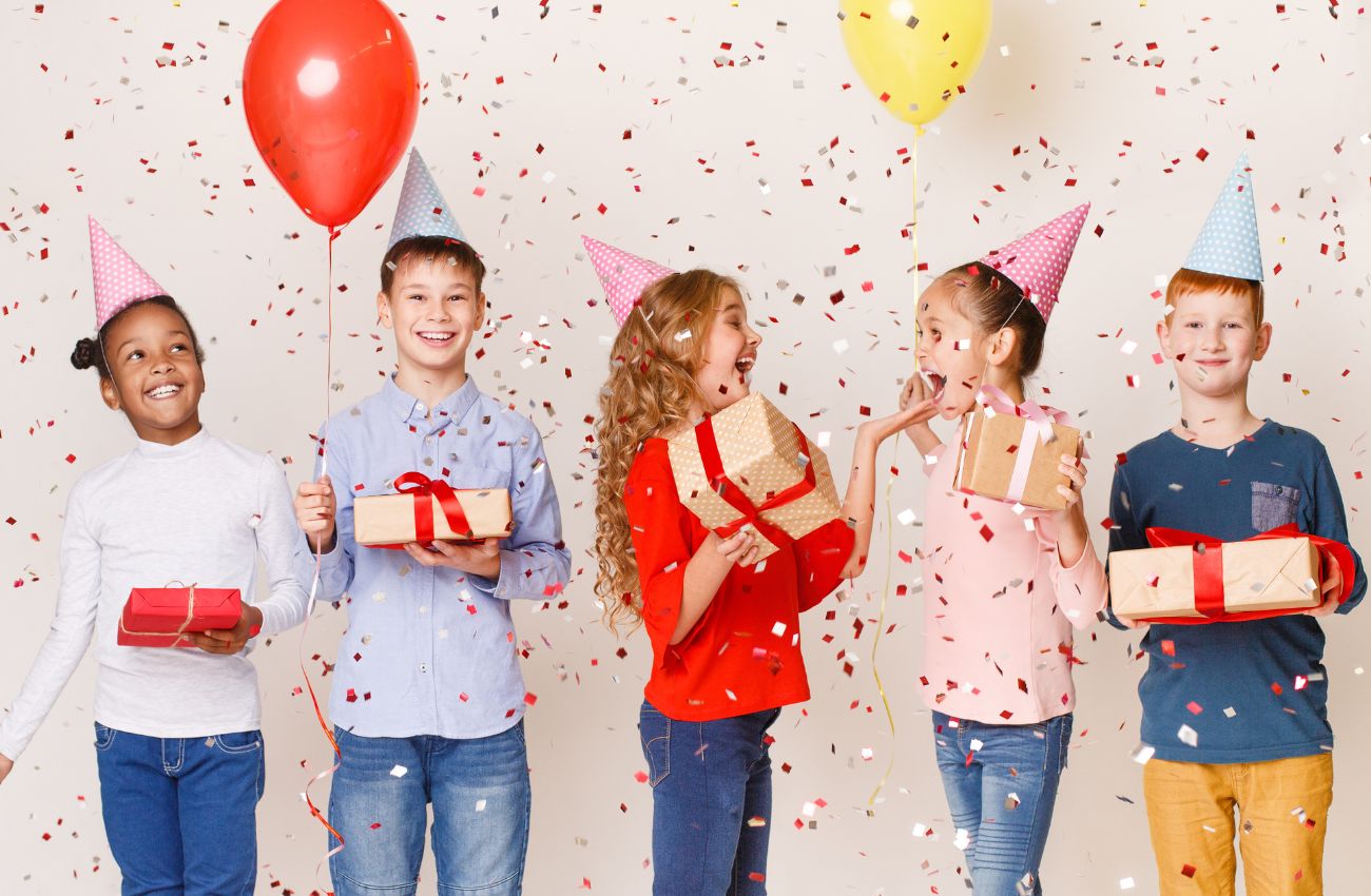 Five kids in party hats hold presents at a birthday party.