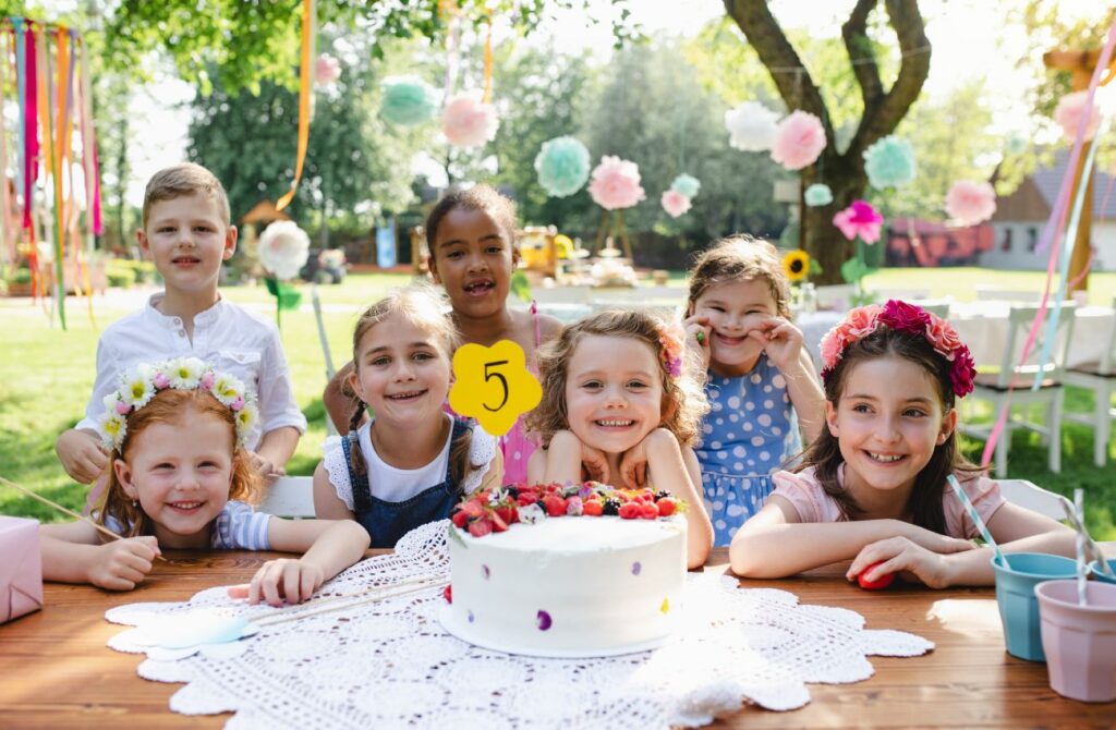 Seven kids smile and sit in front of a white cake outside at a birthday party. 