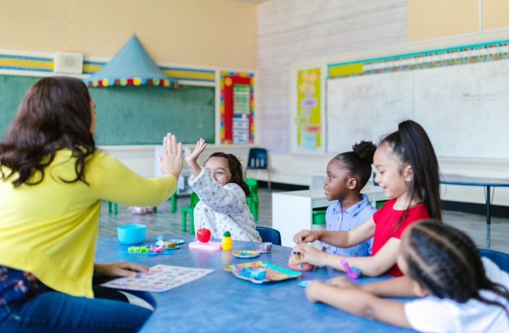 A teacher give a young student a high five in the classroom while three other students watch. 