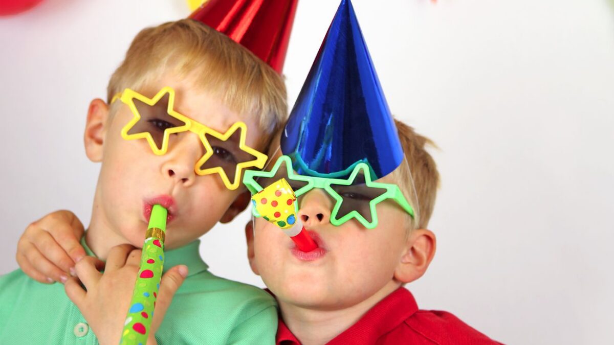 Two boys wear party hats and star sunglasses, and blow party favors.