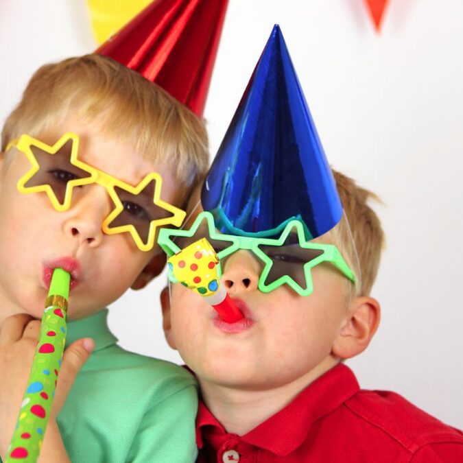 Two boys wear party hats and star sunglasses, and blow party favors.