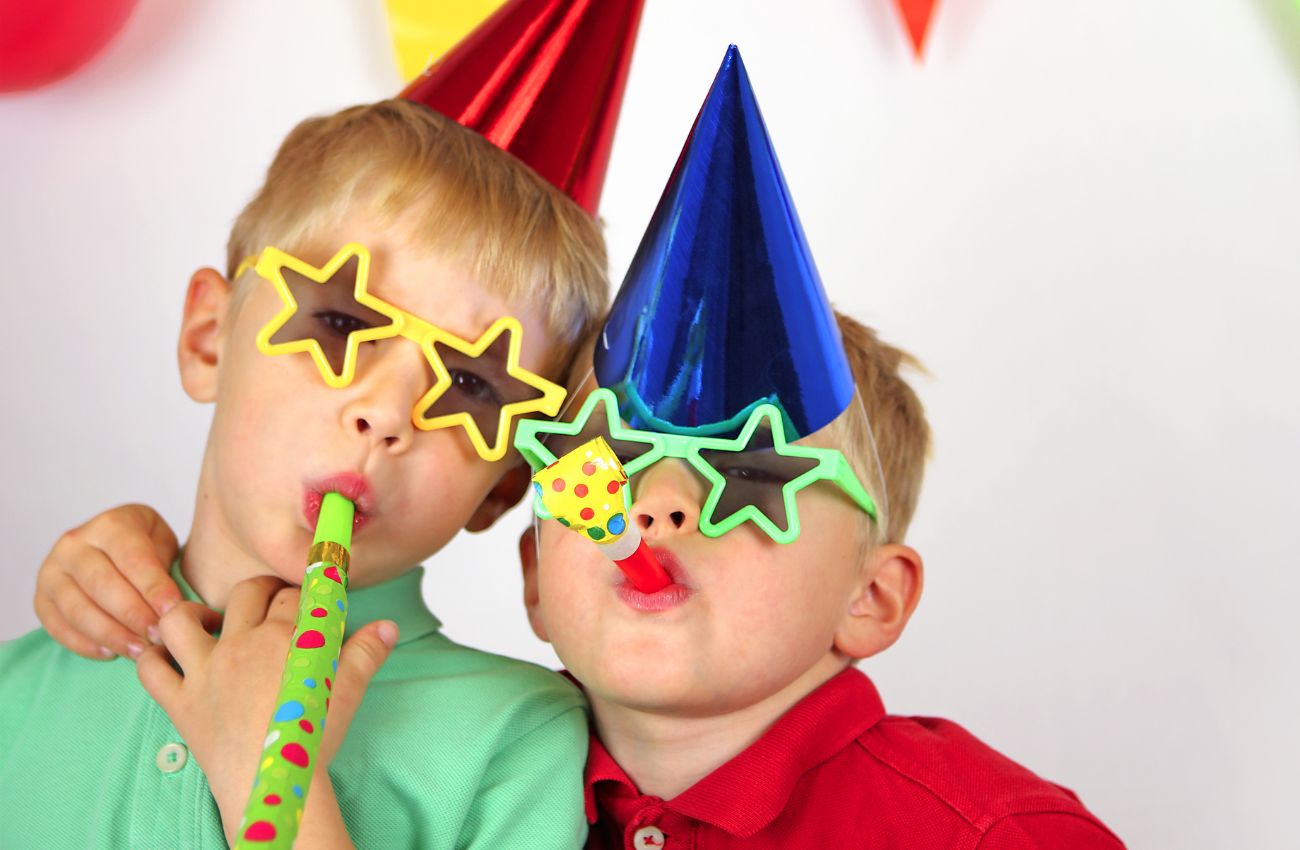 Two boys wear party hats and star sunglasses, and blow party favors.